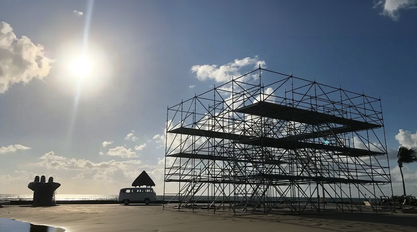 Estructura Layher en proceso de montaje frente al mar, con plataformas modulares, escaleras de acceso y riggers trabajando en la instalación para evento al aire libre.