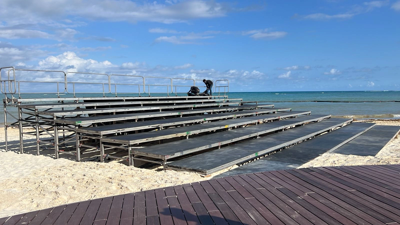 Gradas modulares de andamiaje y madera instaladas en la zona de público de un festival al aire libre, con el escenario principal visible al fondo bajo un cielo despejado
