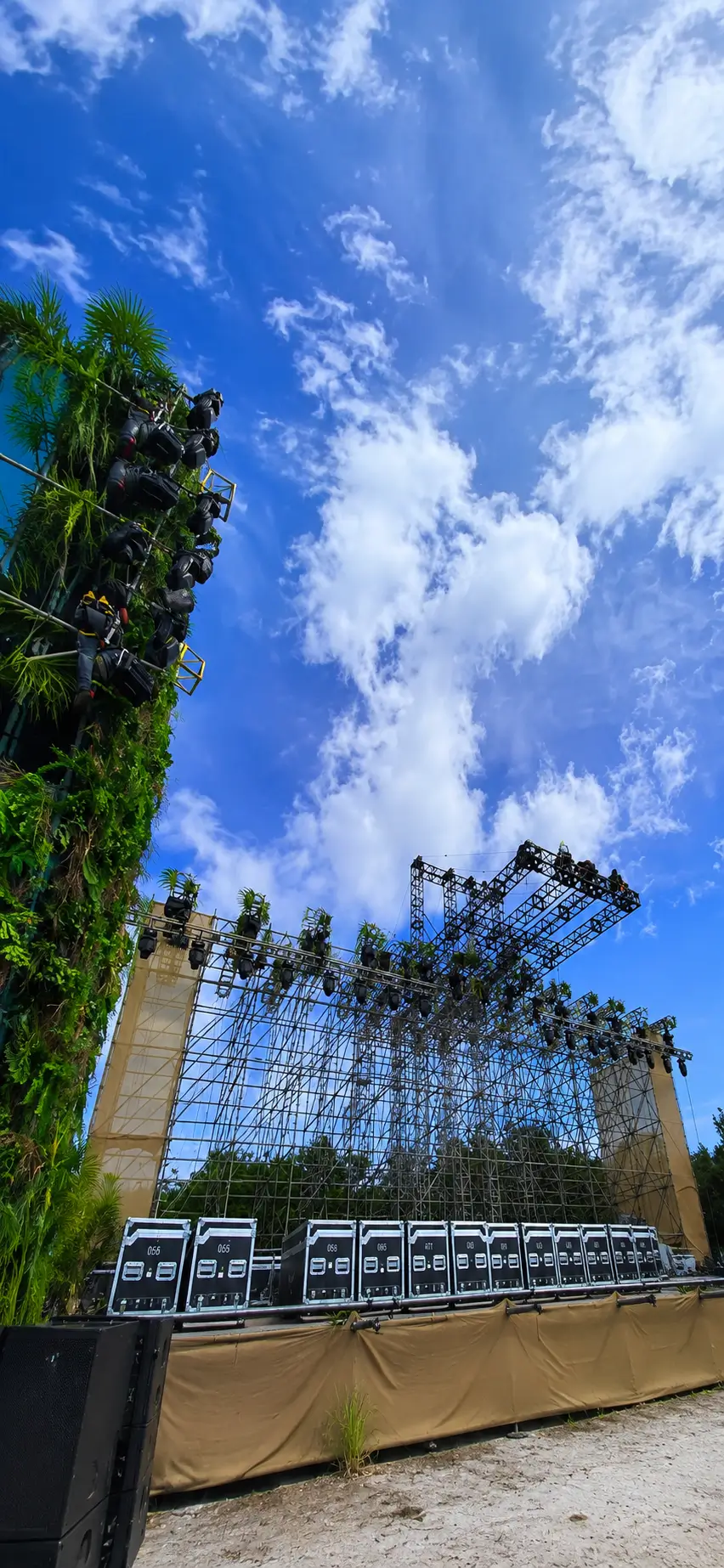 ESCENARIO DE GRAN FORMATO CON ESTRUCTURA DE RIGGING Y TORRES ESCÉNICAS DECORADAS CON VEGETACIÓN, INSTALADO PARA FESTIVAL AL AIRE LIBRE EN ENTORNO TROPICAL.