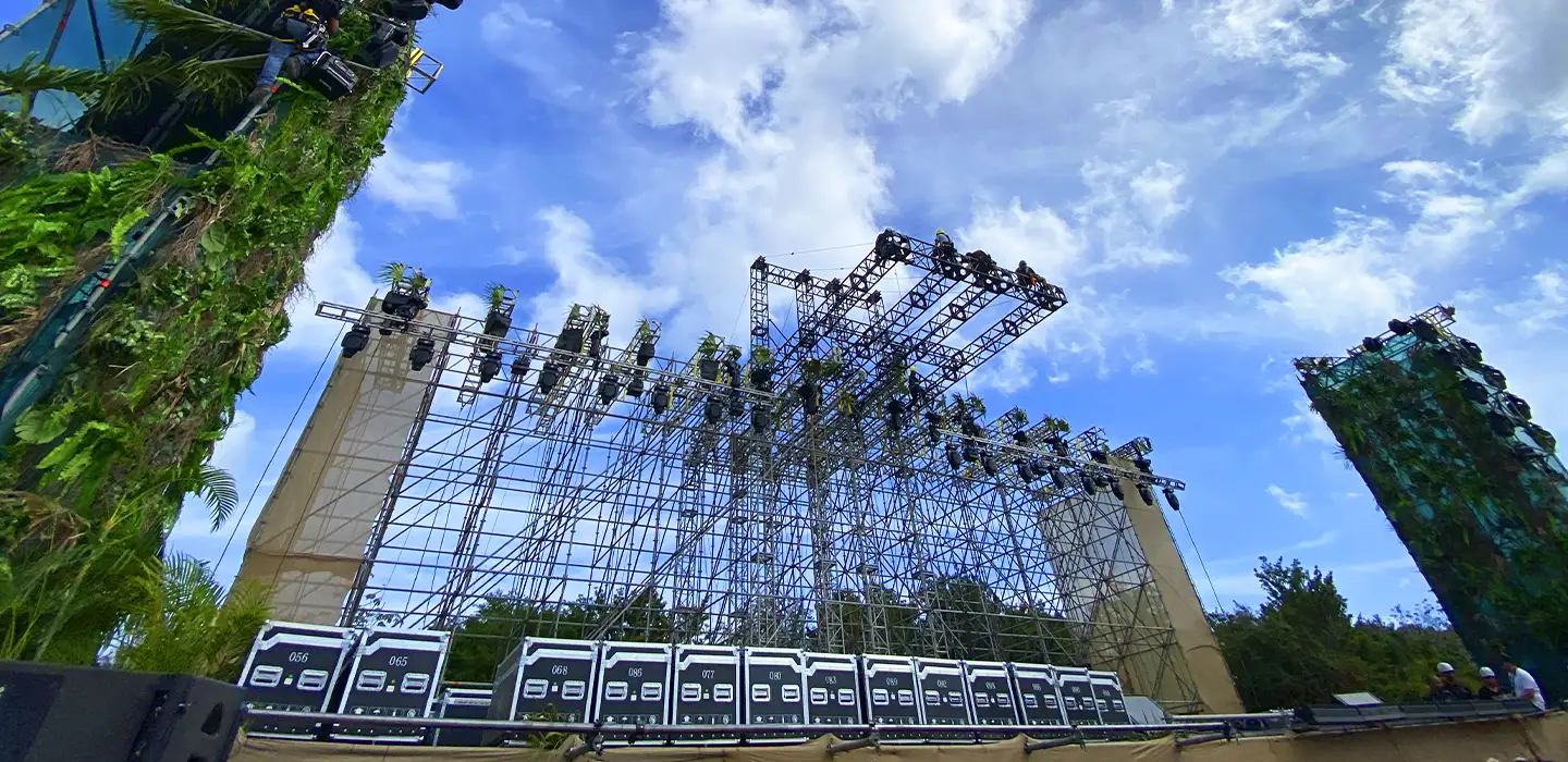 ESCENARIO DE GRAN FORMATO CON ESTRUCTURA DE RIGGING Y TORRES ESCÉNICAS DECORADAS CON VEGETACIÓN, INSTALADO PARA FESTIVAL AL AIRE LIBRE EN ENTORNO TROPICAL.